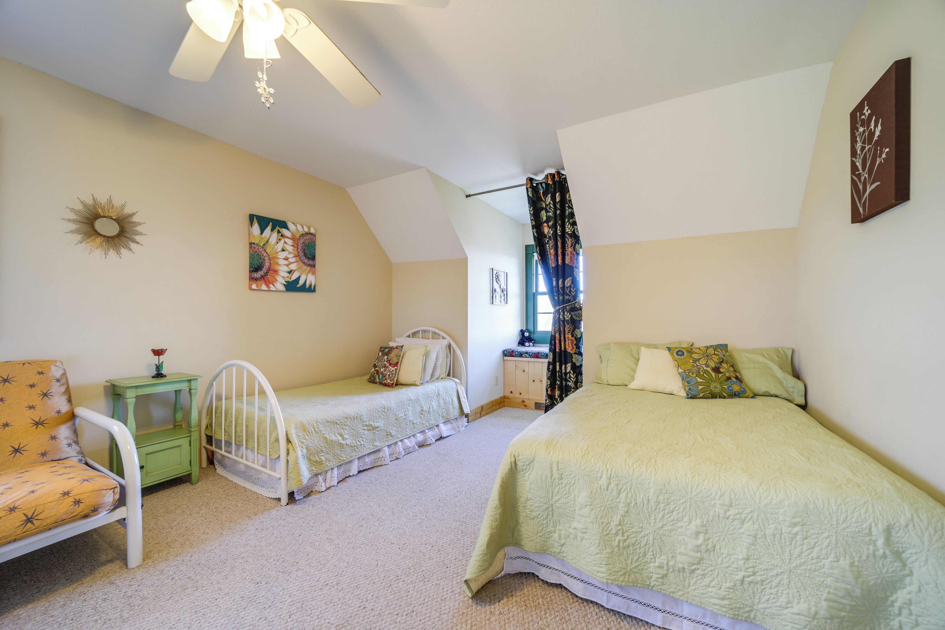 Une chambre avec deux lits simples, une petite table de chevet verte, une chaise blanche et un ventilateur de plafond.