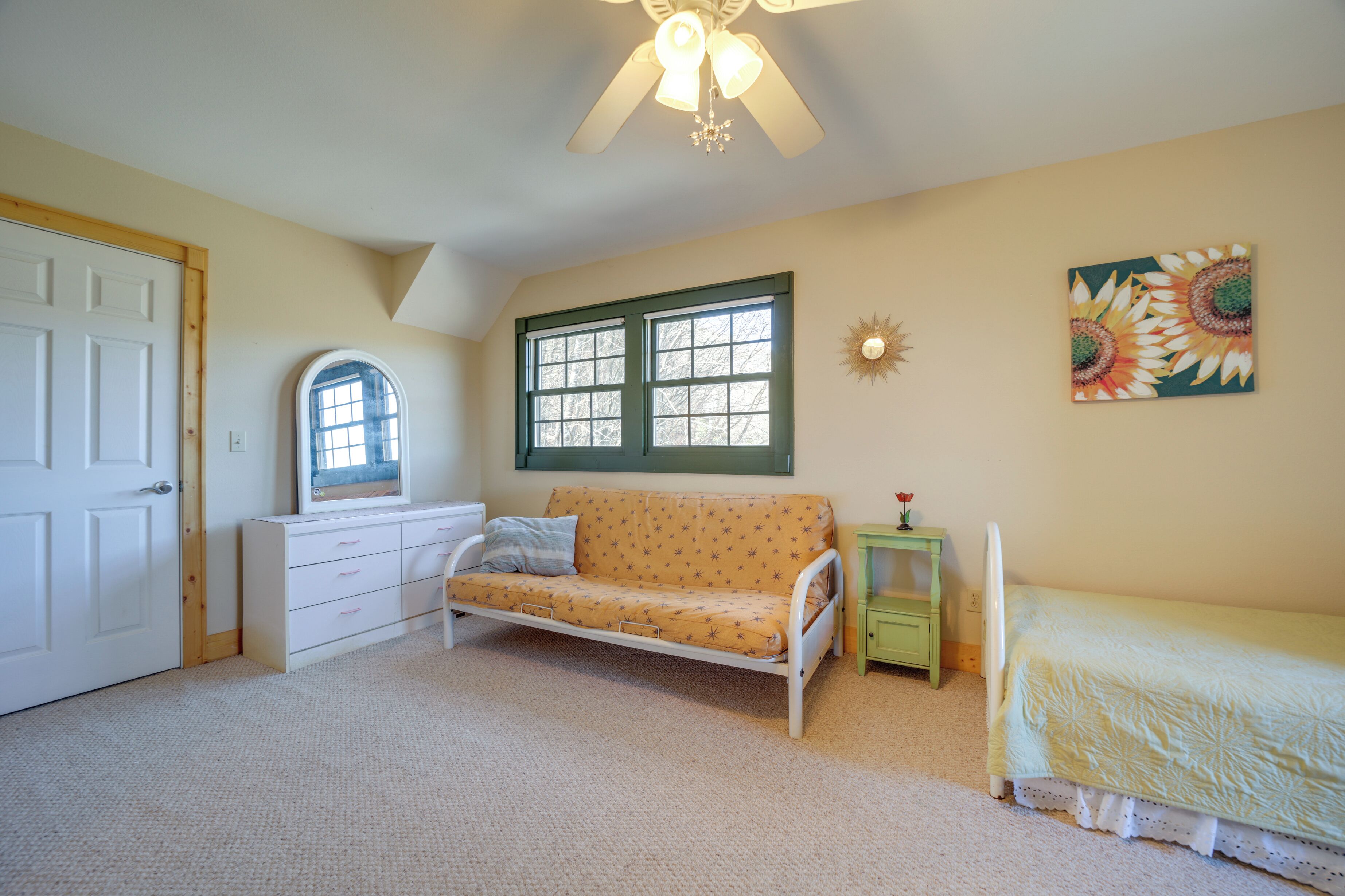 A bedroom with a white bed, a sofa, a window with green shutters, a ceiling fan, and a wall-mounted decorative piece.