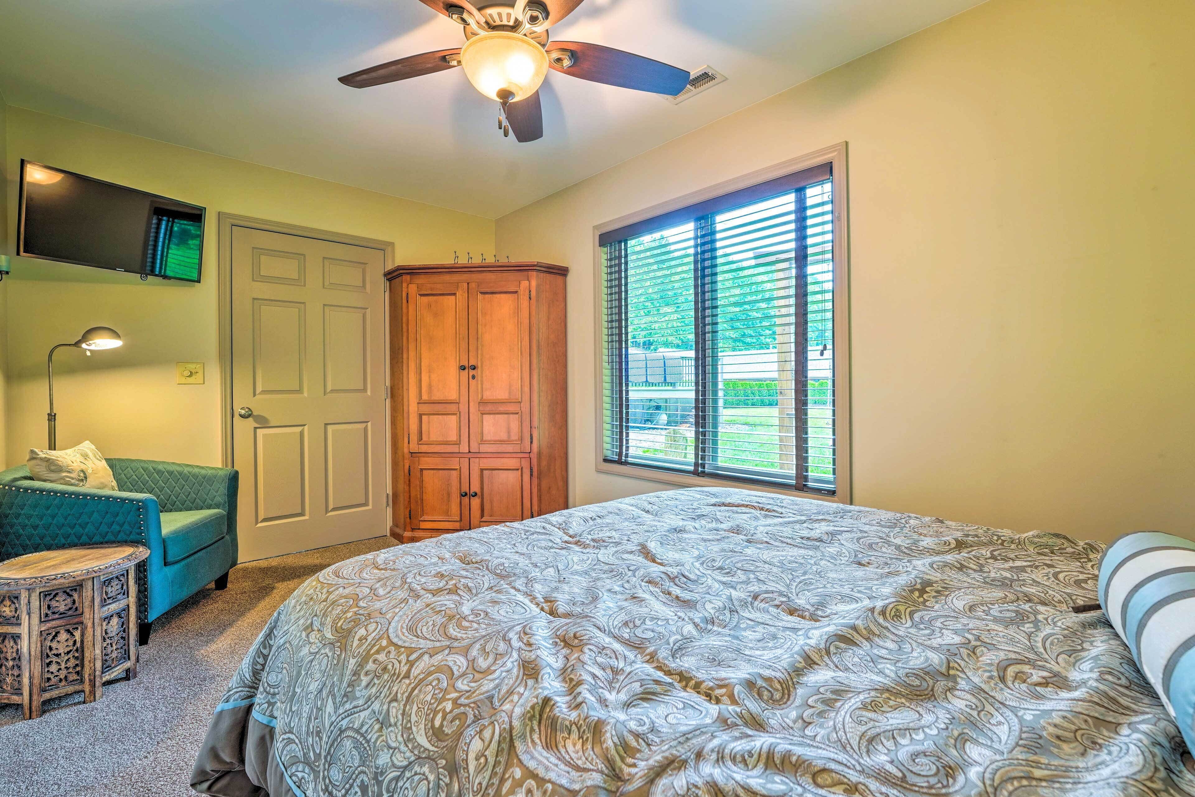 Une chambre avec un grand lit, un ventilateur de plafond, un téléviseur fixé au mur, une armoire en bois, un fauteuil vert et une fenêtre avec des stores.