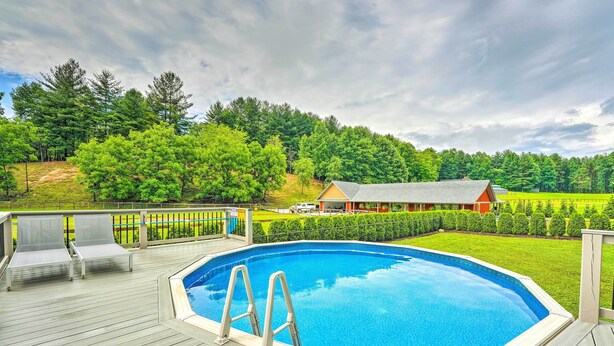 An outdoor swimming pool with a wooden deck, lounge chairs, and a view of a rural landscape with trees and a house.