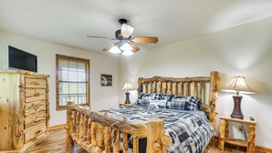 A bedroom with a wooden bed frame, a ceiling fan, a TV, and a window with blinds.