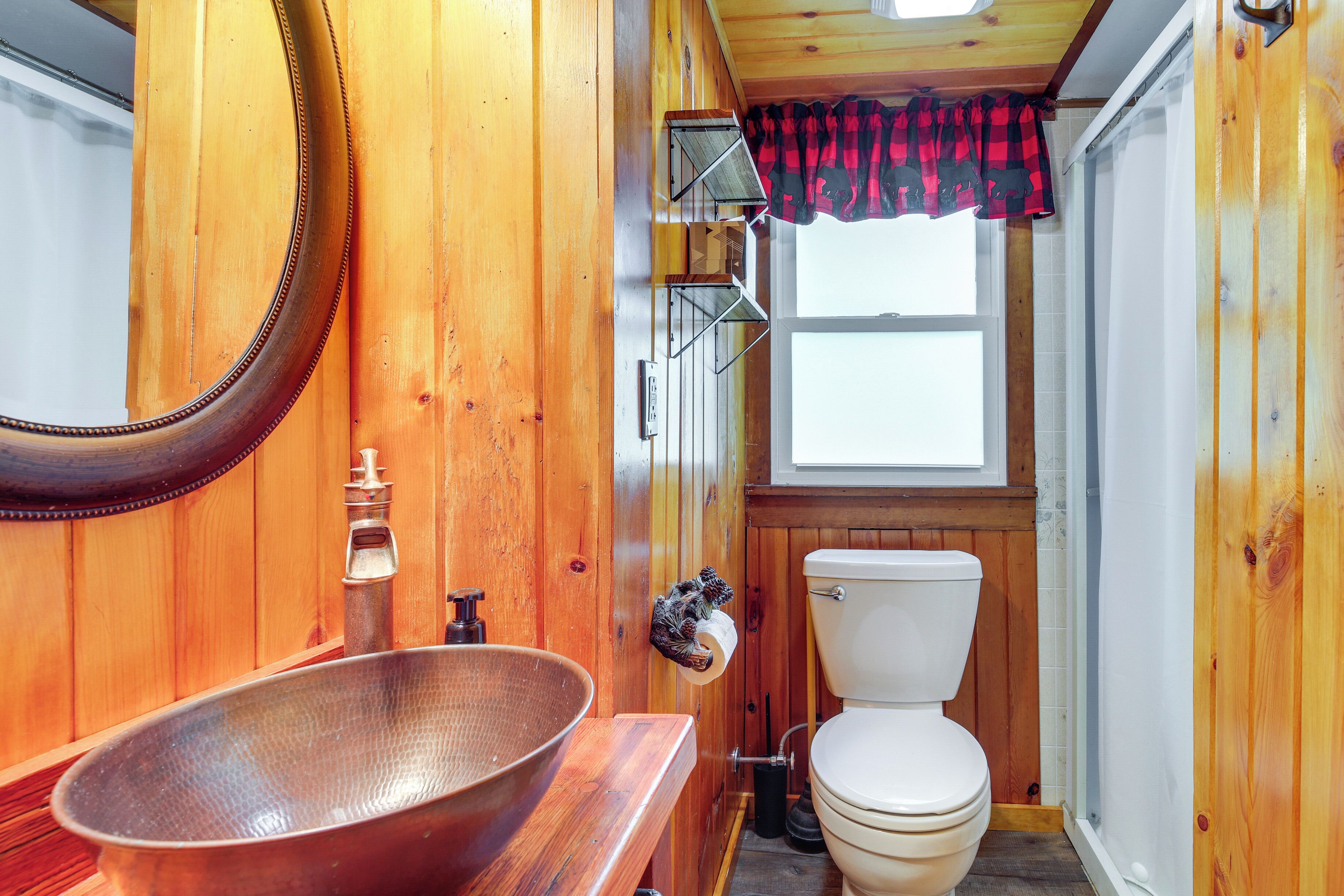 Ein Badezimmer aus Holz mit einem großen, ovalen Waschbecken, einer Toilette und einem Fenster mit einem rot-schwarz karierten Vorhang.