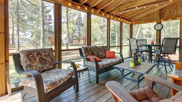 A covered outdoor seating area with wicker furniture, a table, and chairs, overlooking a forested area. - Dock & Lake Access! St Germain Getaway (St Germain)