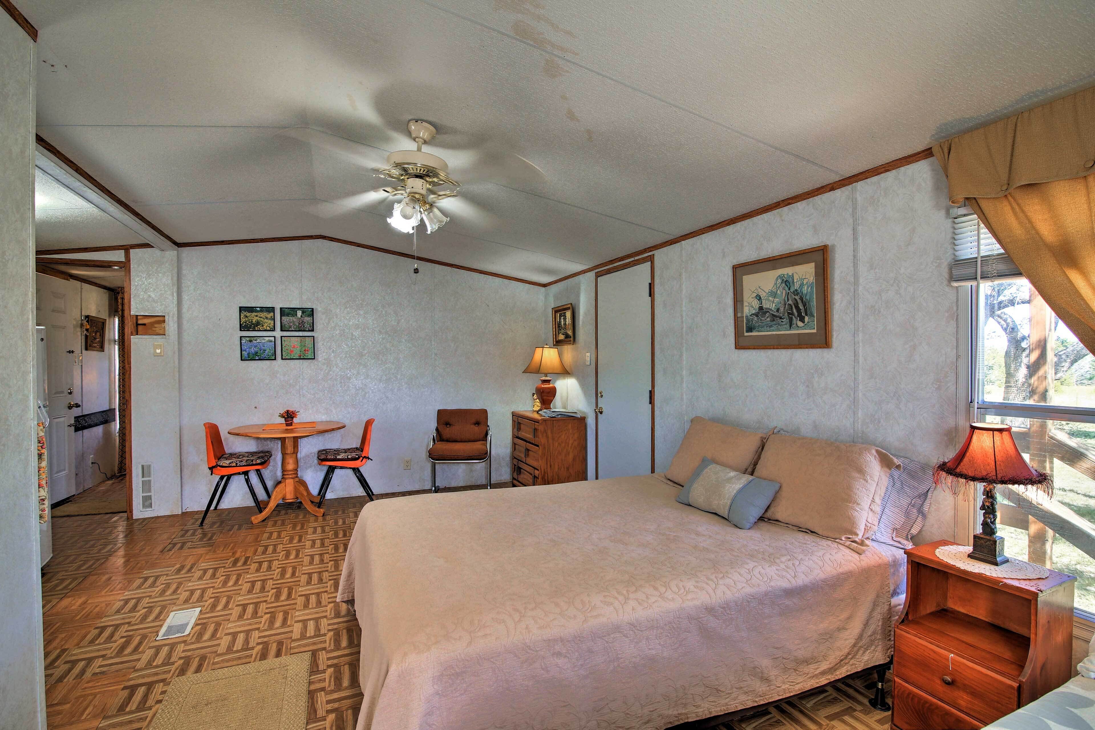 A bedroom with a bed, a desk with chairs, a ceiling fan, a lamp, and a window with curtains.