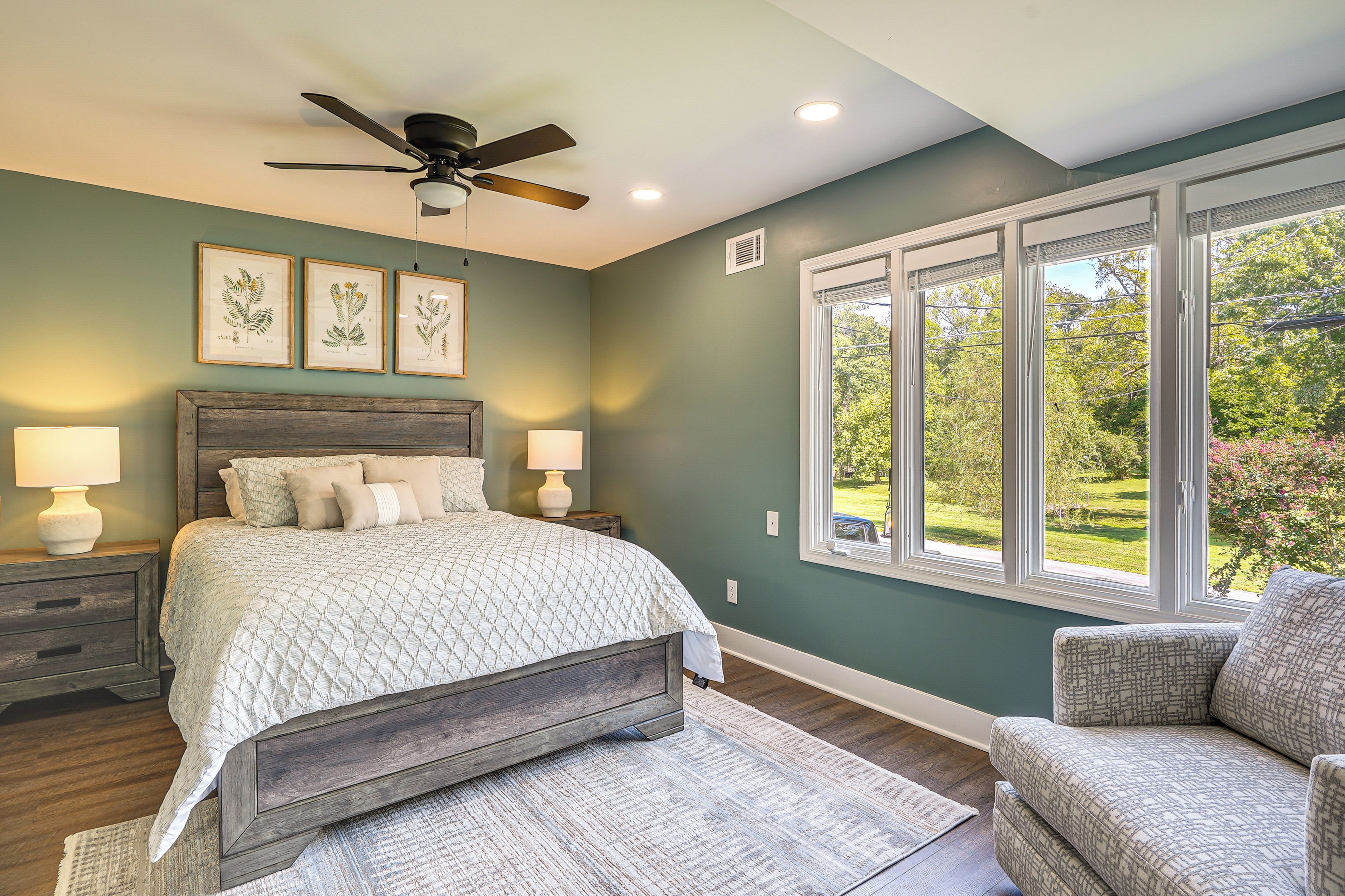 A bedroom with a bed, nightstands, a ceiling fan, framed botanical prints, and a window with a view of greenery.
