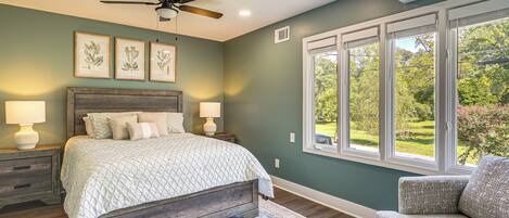 A bedroom with a bed, nightstands, a ceiling fan, framed botanical prints, and a window with a view of greenery.