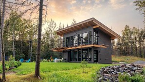 Een modern huis met twee verdiepingen en een groot raam, omringd door een bos.