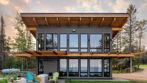 Een modern huis met twee verdiepingen, grote ramen, een overdekt terras en uitzicht op het omliggende bos.