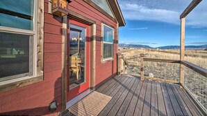 A red wooden house with a deck, large windows, and a view of a grassy field and mountains. - Romantic Gem w/ Mtn Views! Day Trip to Yellowstone (Livingston)