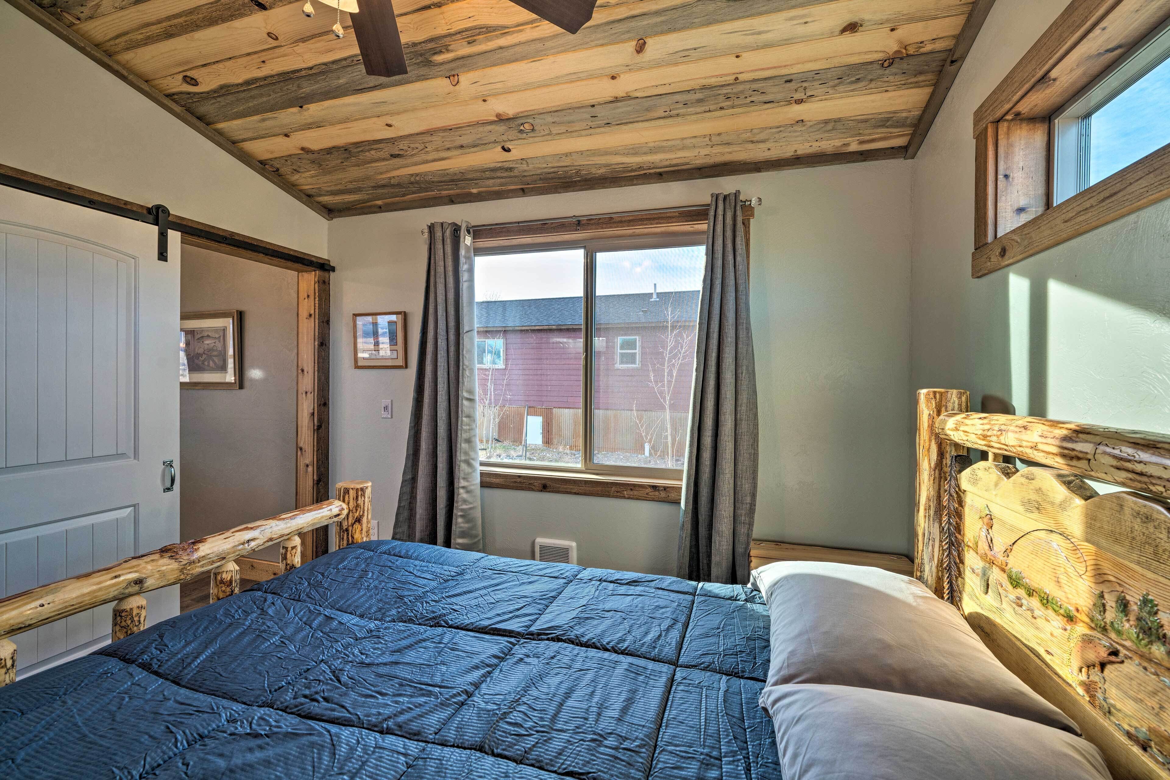 A bedroom with a wooden ceiling, a large bed with a blue quilt, a wooden headboard, and a window with curtains.