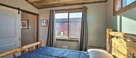 A bedroom with a wooden ceiling, a large bed with a blue quilt, a wooden headboard, and a window with curtains.