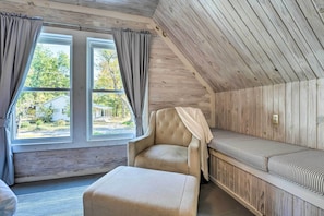 A cozy room with a wooden ceiling, a cushioned bench, a cushioned chair, and a large window with curtains. - Unique Loft Cabin: Fire Pit & Fishing Access! (Bells)