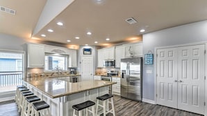 A modern kitchen with a large island, white cabinetry, stainless steel appliances, and a wooden floor. - Group Getaway w/ Deck, Steps to Crystal Beach! (Port Bolivar)