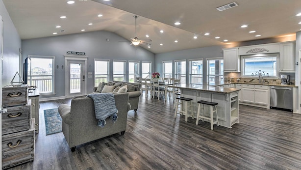 A spacious living area with a gray sofa, a dining table, and a kitchen with white cabinets and a bar. - Group Getaway w/ Deck, Steps to Crystal Beach! (Port Bolivar)