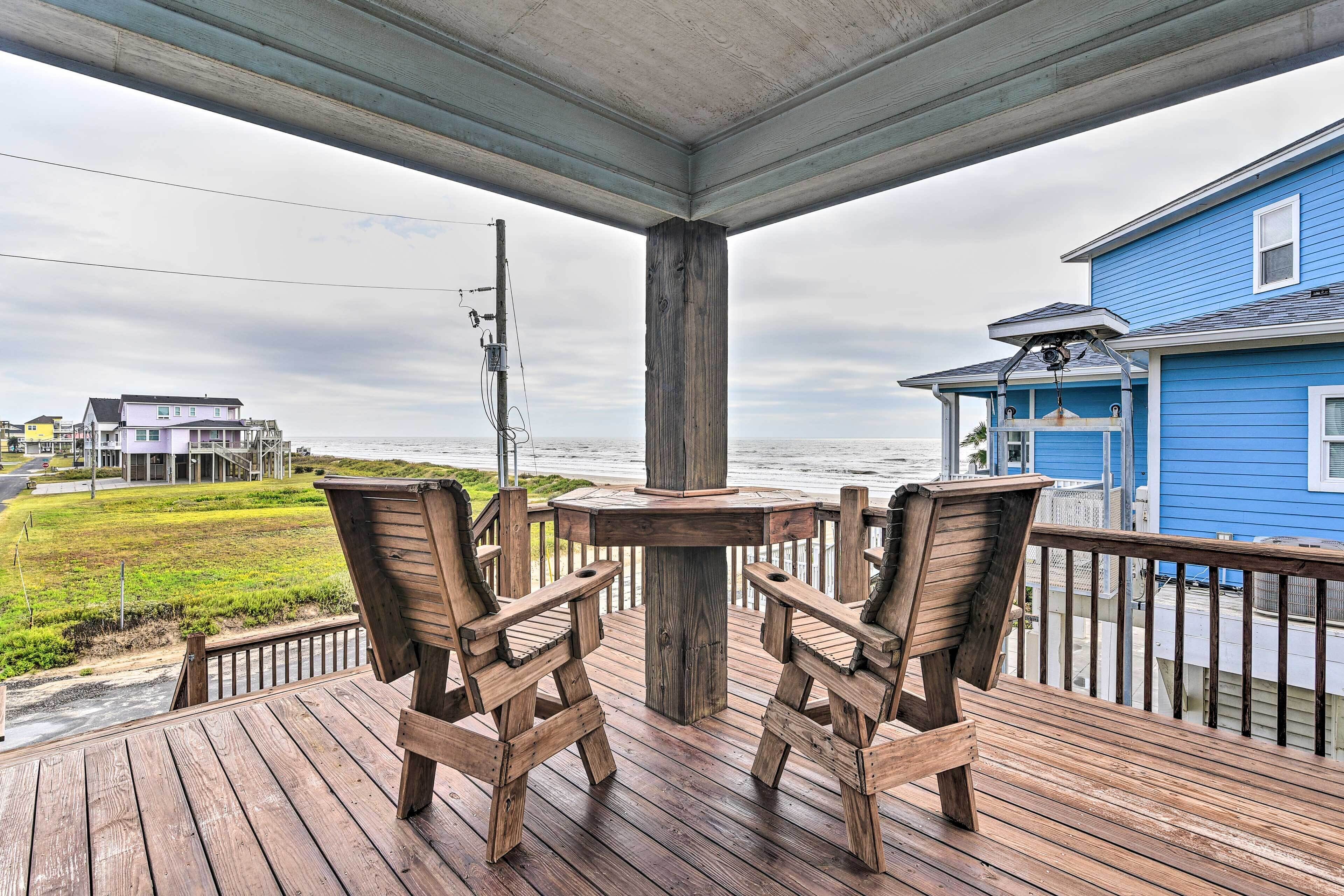 A wooden deck with two chairs and a table, overlooking a beach and a blue house.