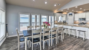 A modern kitchen with white cabinetry, a central island, and a dining area with a wooden table and chairs. - Group Getaway w/ Deck, Steps to Crystal Beach! (Port Bolivar)