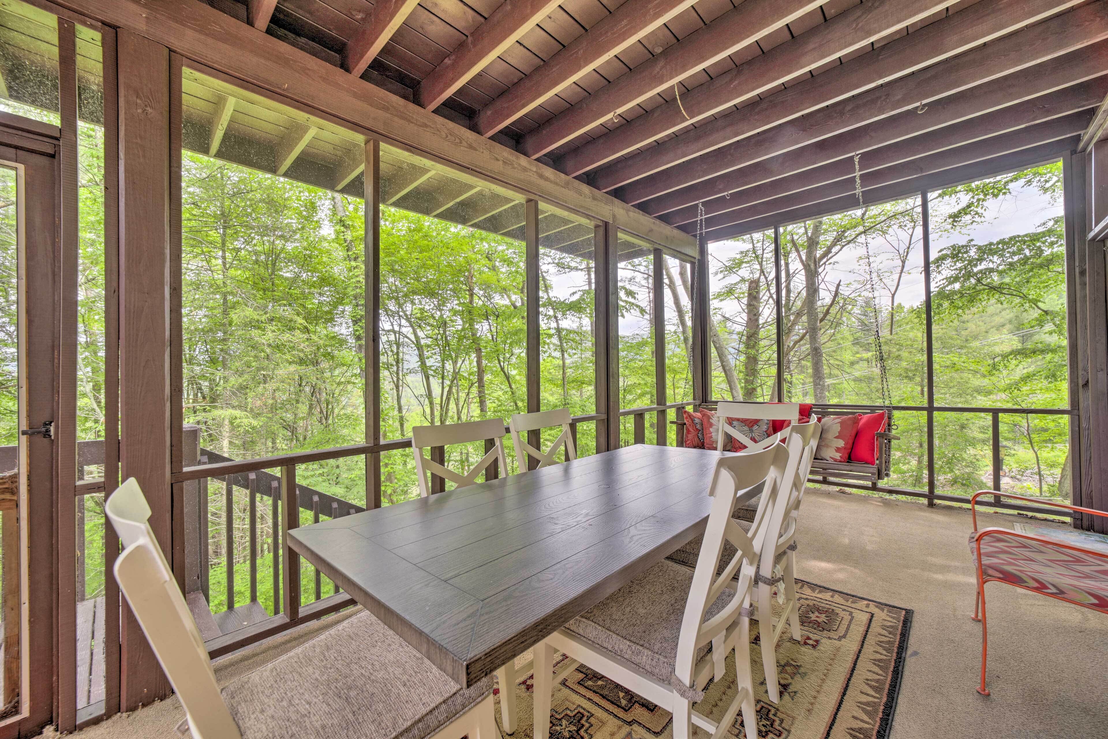A covered outdoor dining area with a wooden table and white chairs, overlooking a forested area.