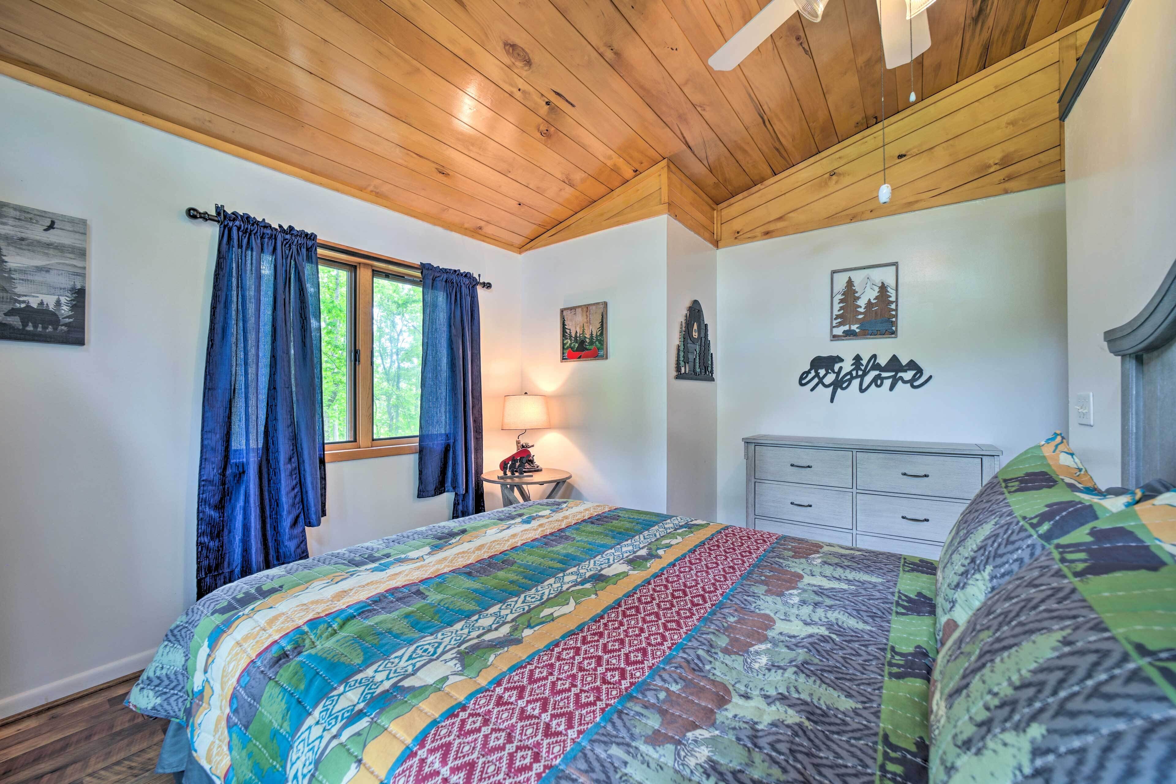 A bedroom with a wooden ceiling, a bed with a colorful quilt, a nightstand with a lamp, and a dresser.