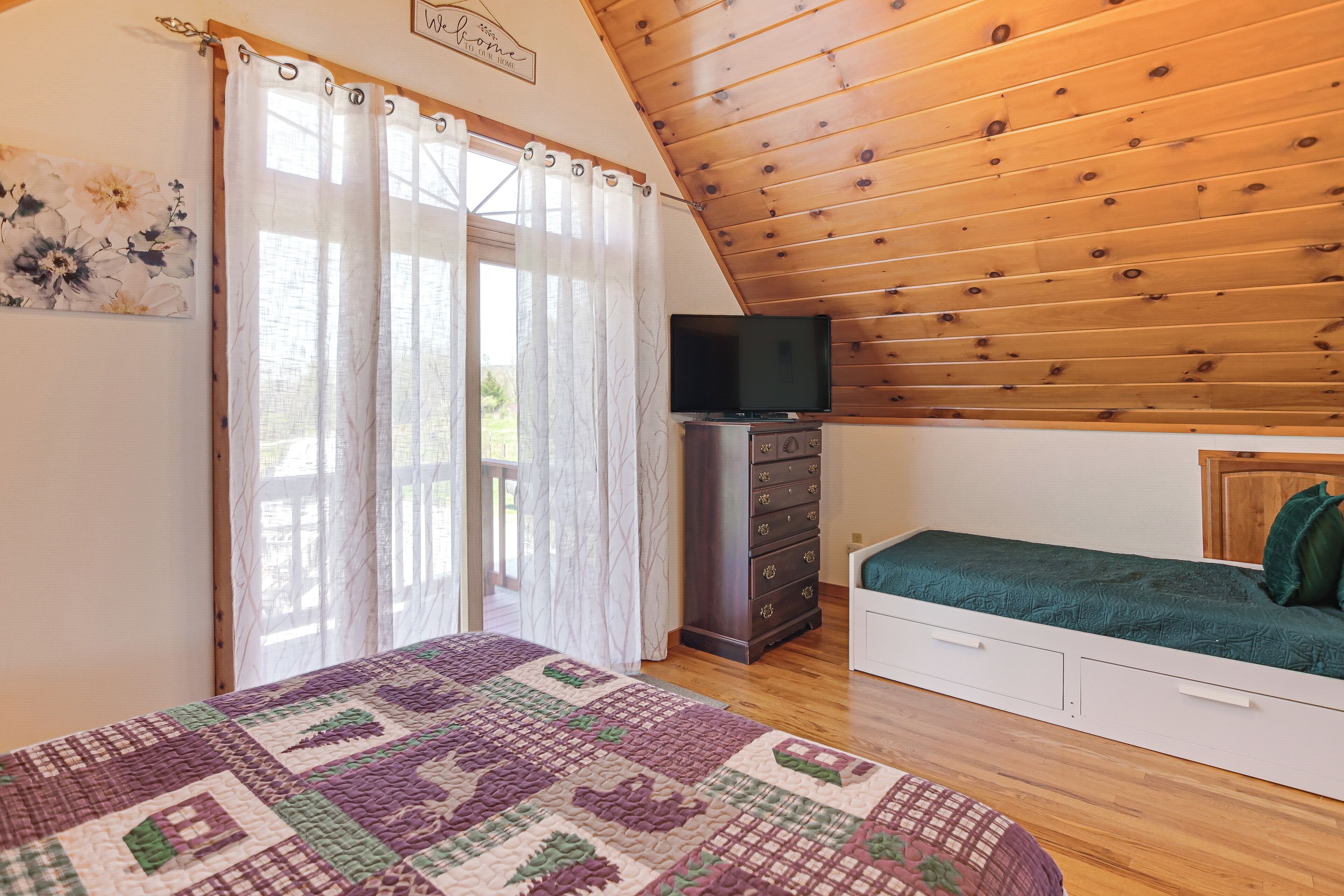 A bedroom with a wooden ceiling, a bed with a green blanket, a dresser, and a TV on a stand.