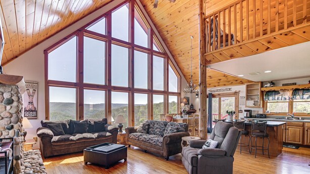 A spacious living room with large windows, a stone fireplace, and a kitchen area. - West Virginia Cabin Near Snowshoe Mountain Resort (Slaty Fork)
