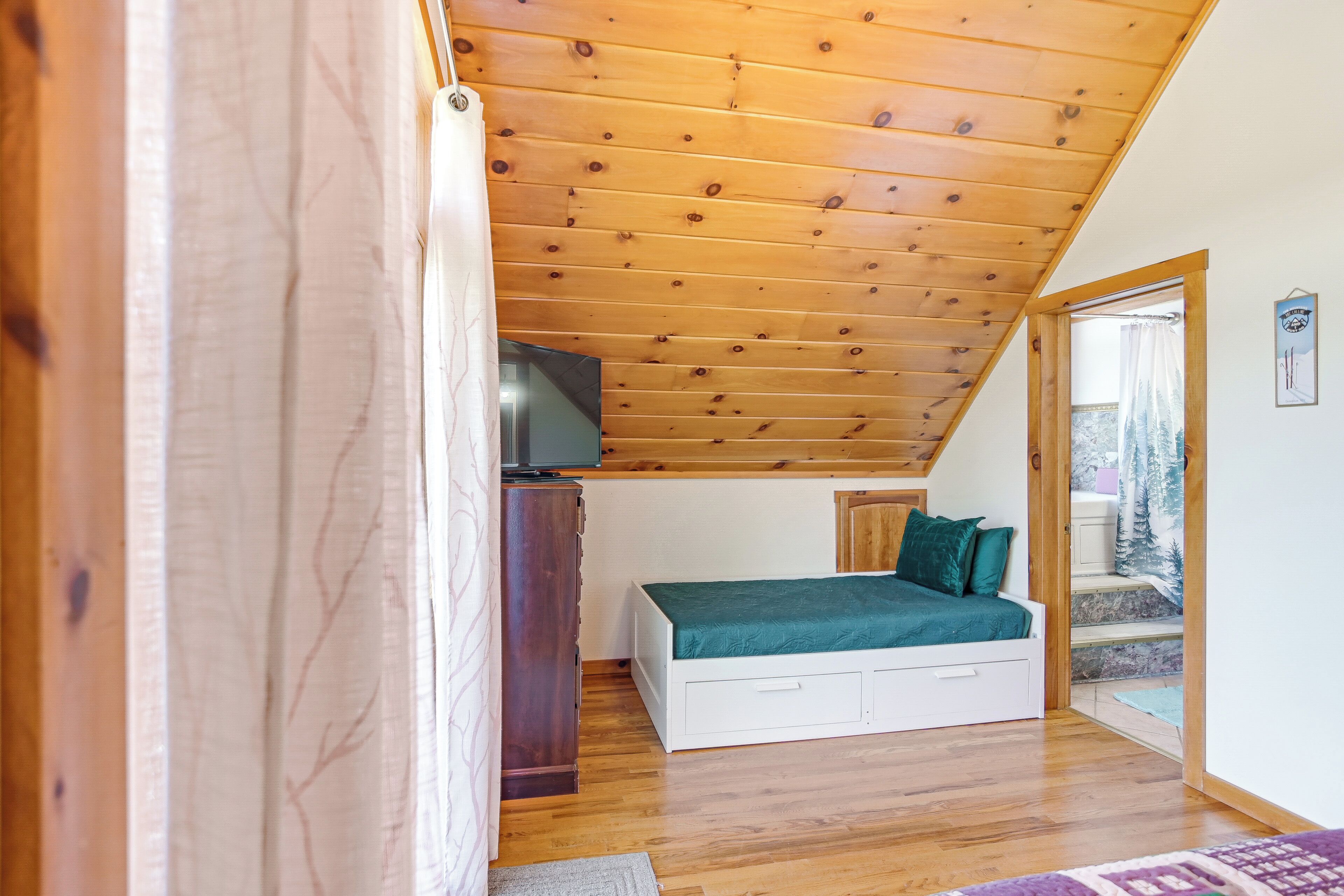 A bedroom with a sloped ceiling, a bed with a green cover, a wooden dresser, and a television on top.