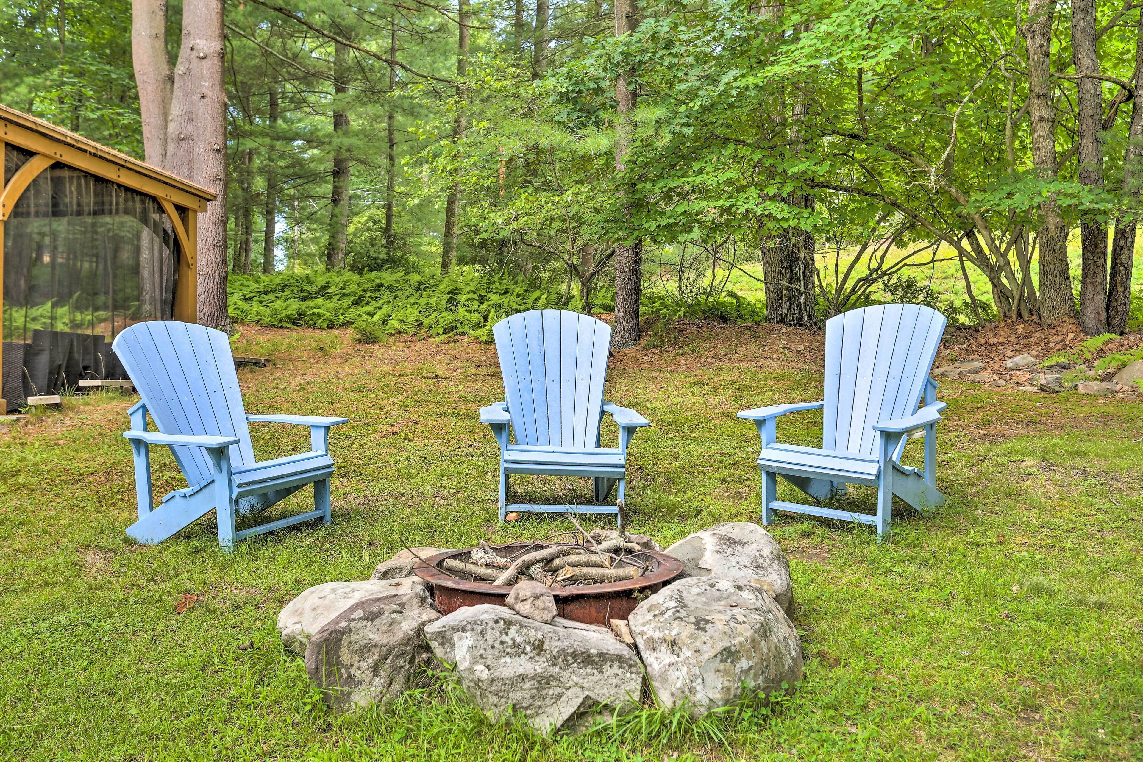 Three blue Adirondack chairs arranged around a fire pit in a forested area with a wooden structure in the background.
