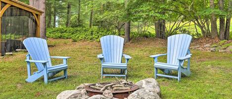Three blue Adirondack chairs arranged around a fire pit in a forested area with a wooden structure in the background.