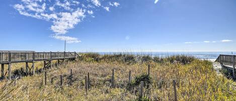 Un paseo marítimo de madera que lleva a una playa con pasto seco y un cielo despejado.