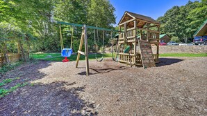 A wooden play structure with a slide and swings, surrounded by trees and a clear sky. - In Golf Community w/ Pool Access: Newland Cabin (Newland)
