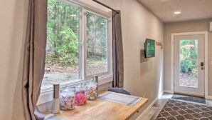 Una habitación con una mesa de madera, una ventana con cortinas, una puerta y vistas a árboles al exterior.