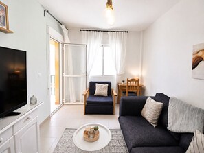 A living room with a sofa, a chair, a TV, a table with a lamp, and a window with curtains. - Apartamento El Calón Playa - Lateral Luz (El Calón)