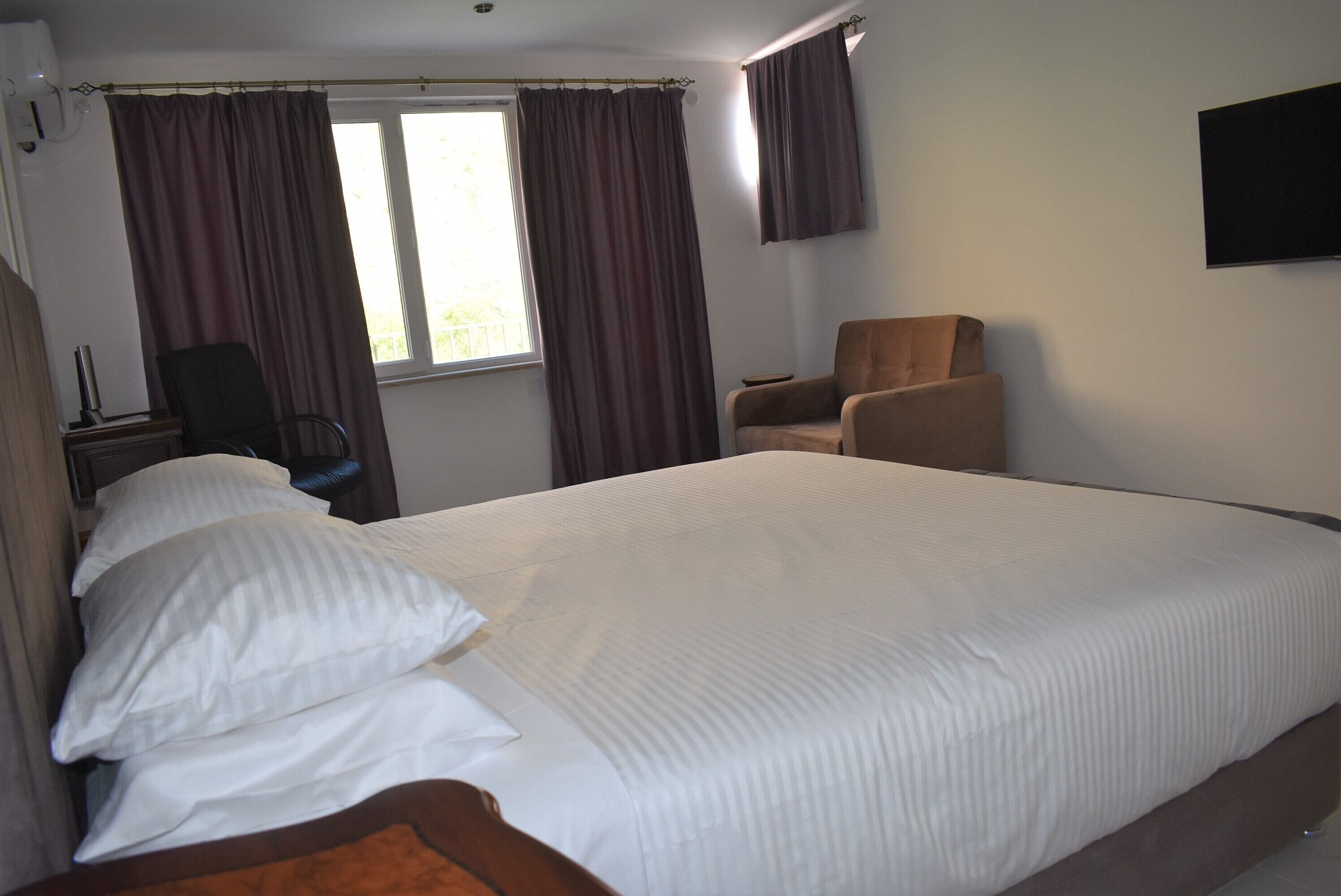 A hotel room with a large bed, two chairs, a desk with a lamp, and a window with curtains.