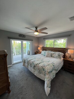 A bedroom with a large bed, a ceiling fan, a nightstand with a lamp, and a dresser.