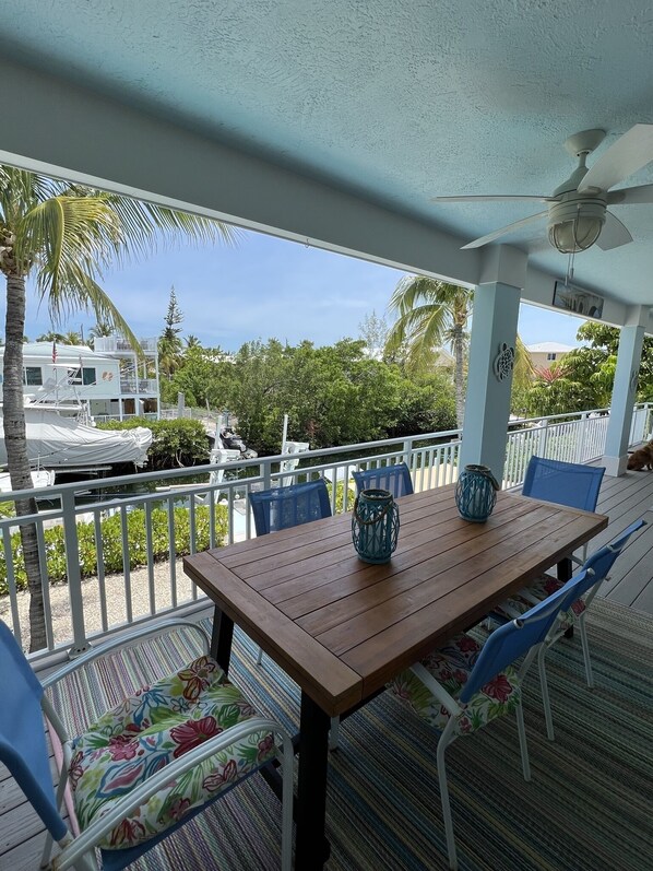A covered outdoor dining area with a wooden table and blue chairs, surrounded by a white railing and palm trees.