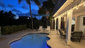 Una piscina con bordo ondulato, circondata da lettini prendisole e una zona patio coperta con luci decorative.