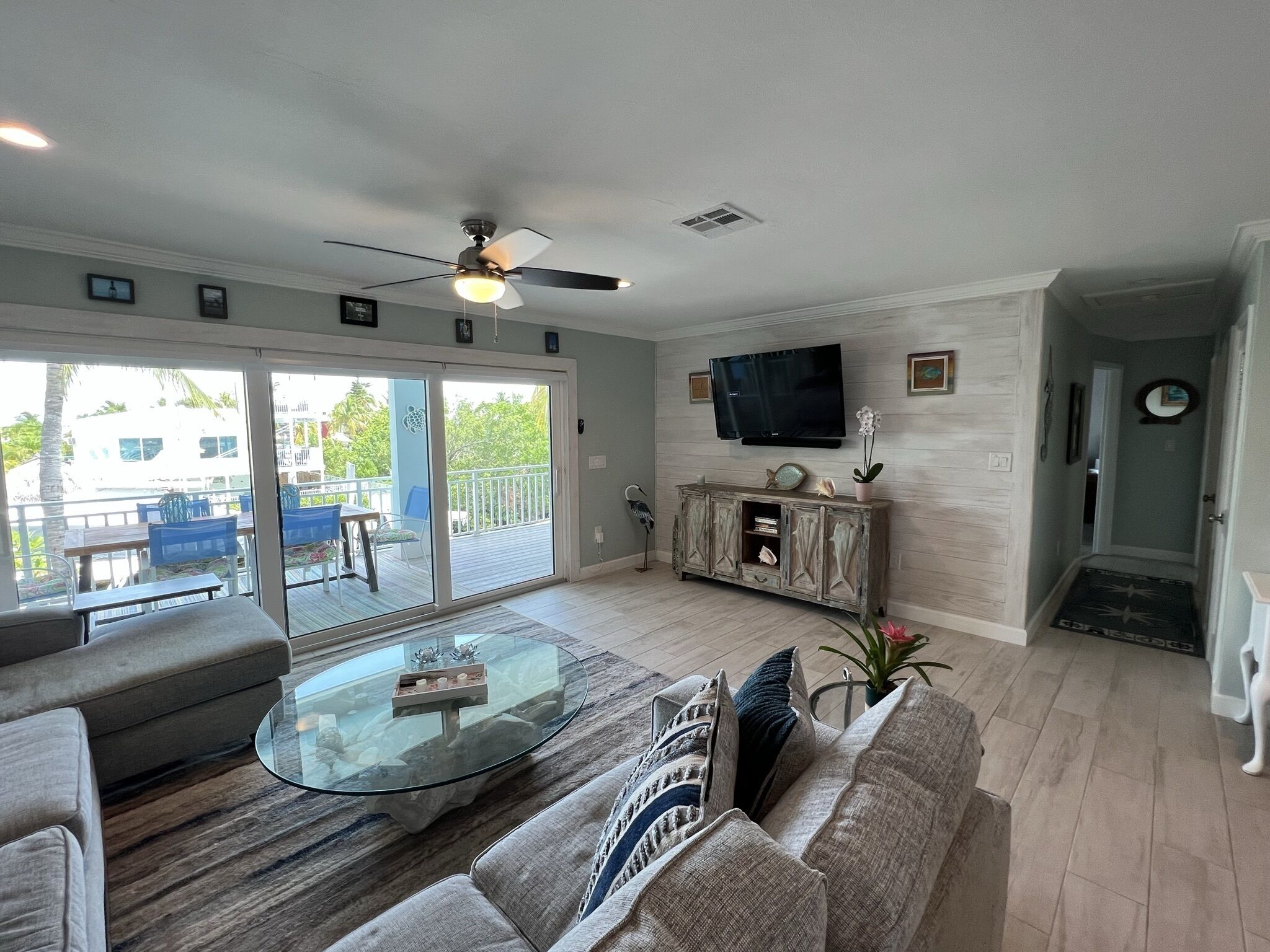 A living room with a glass coffee table, a sofa, a TV mounted on the wall, and a sliding glass door leading to an outdoor area with blue chairs.