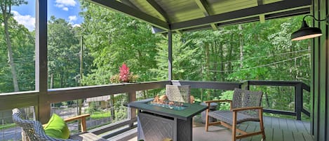 A covered outdoor deck with wicker furniture, a table with a fire feature, and a view of a forested area.