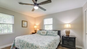 Un dormitorio con una cama, dos mesitas de noche, un ventilador de techo y dos ventanas con persianas.