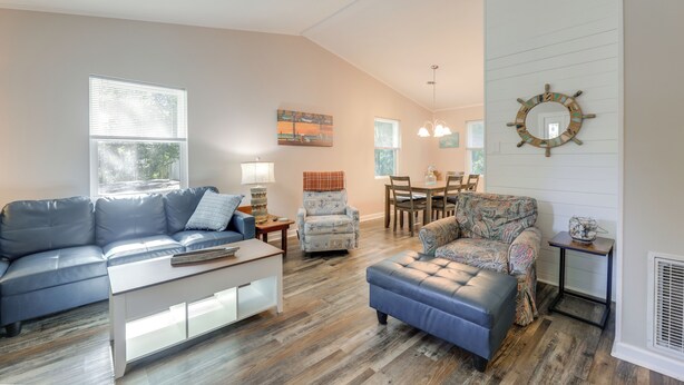 Una sala de estar con un sofá azul, un sillón estampado y un reposapiés, piso de madera y un ventilador de techo.