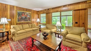 A living room with wooden paneled walls, two beige sofas, a wooden coffee table, and a red patterned rug. - Near Hiking: Home w/ Trout Stream in Bakersville! (Bakersville)