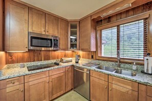 A kitchen with wooden cabinets, a stainless steel microwave, a built-in oven, a granite countertop, a double sink, and a window with blinds. - Near Hiking: Home w/ Trout Stream in Bakersville! (Bakersville)