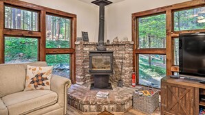 Ein gemütliches Wohnzimmer mit Kamin aus Stein, großen Fenstern mit Blick auf den Wald, einer bequemen Couch und einem Fernseher.