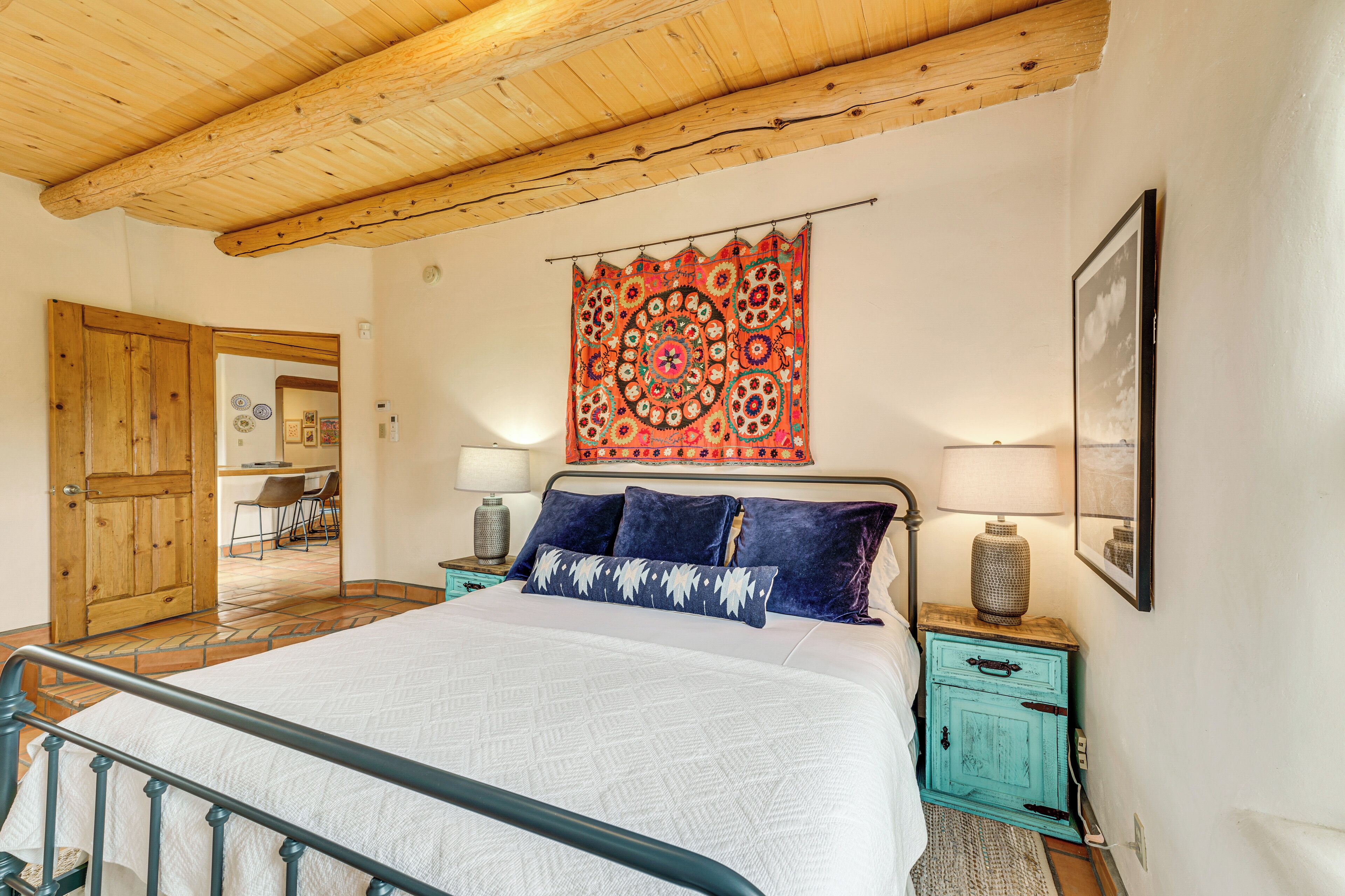 Quarto com teto de madeira, uma cama grande com roupa de cama branca, almofadas azuis e uma tapeçaria colorida na parede.