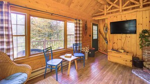 A wooden cabin interior with a dining area, two blue chairs, a wicker sofa, a television mounted on the wall, and large windows offering a view of the outdoors. - Andover Cabin Retreat w/ Hot Tub & Fireplace! (Chester)