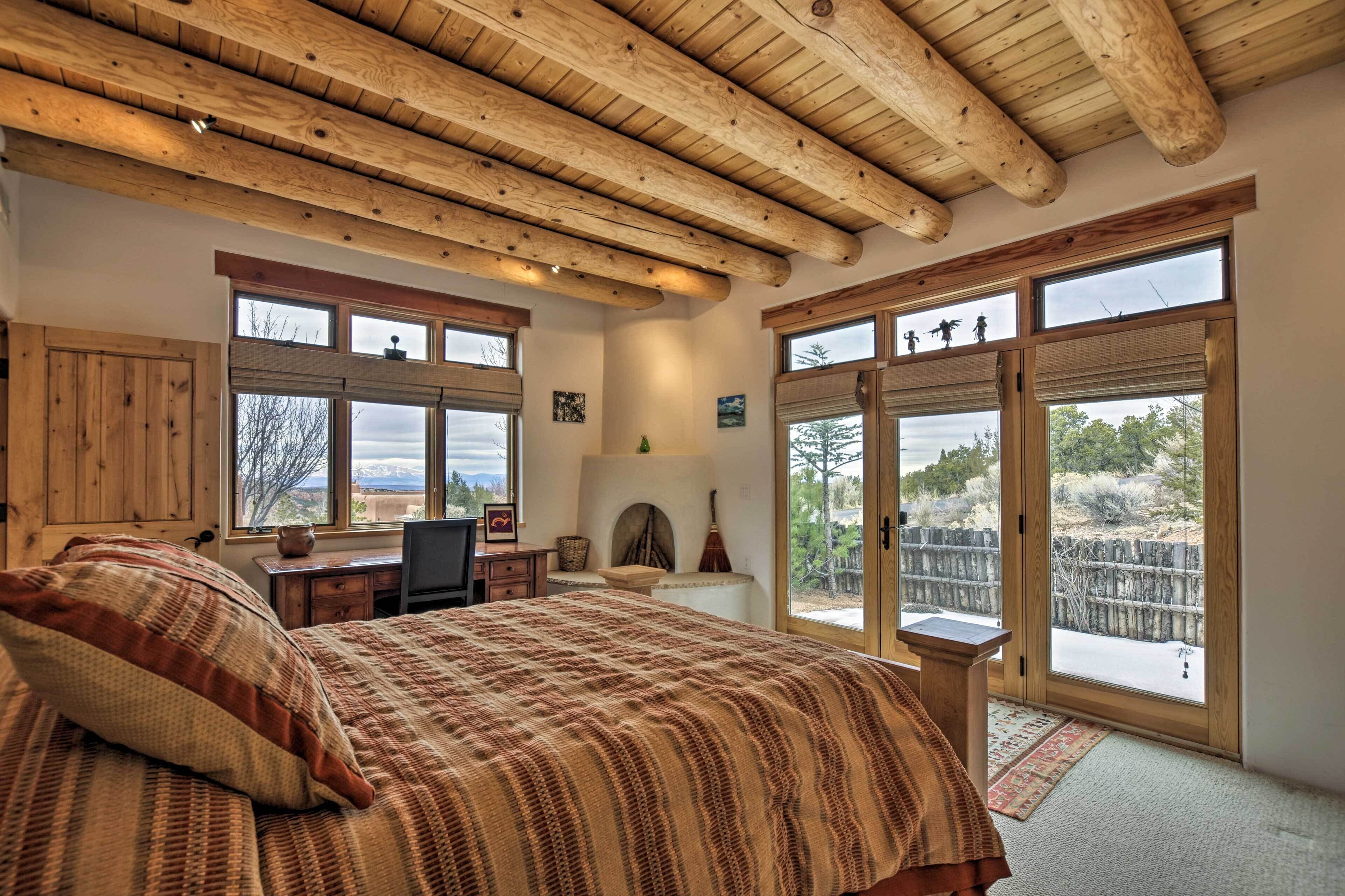 A bedroom with a large bed, wooden ceiling beams, a desk, and a view of the outdoors.
