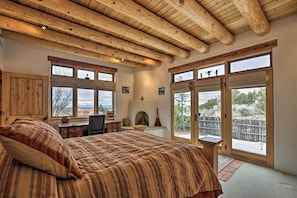 A bedroom with a large bed, wooden ceiling beams, a desk, and a view of the outdoors.