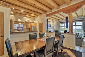A dining area with a wooden table and chairs, a kitchen with wooden cabinets, and a view through large windows.