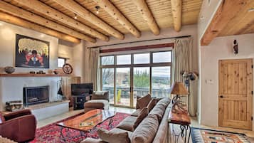 A cozy living room with a fireplace, large windows, and a wooden ceiling.