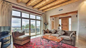 A spacious living room with a large window, a wooden ceiling, a sofa set, and a TV.