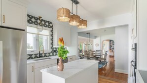 A modern kitchen with white cabinetry, a central island, and a dining area with a wooden table and chairs. - Hot Tub + Private Beach: Waterfront Urbanna Escape (Urbanna)